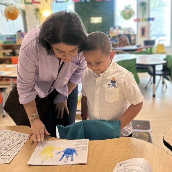 Teacher guides a young student as they review artwork at a classroom table during an ECC Open House.