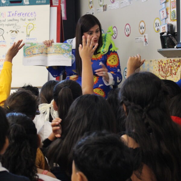 Dresses as Ms. Frizzle, Chief Academic Officer, Dr. Nunez reads a colorful picture book aloud to a classroom of elementary school students during Cityscape Reads while children sit closely together and raise their hands.
