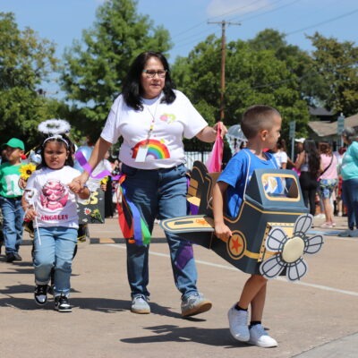 Early Childhood Center students in costumes walk outdoors during the Letter Day Parade; one child wears a cardboard airplane, another wears angel wings and a halo, led by a teacher holding their hands.